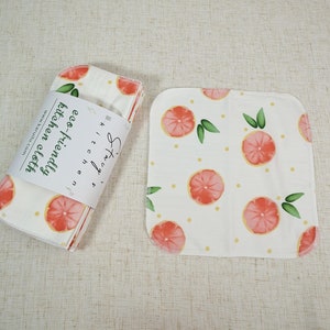 May include: Set of four white kitchen cloths with a grapefruit print. The cloths are folded and stacked, with a label that reads "eco-friendly kitchen cloths".