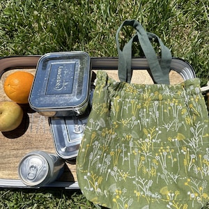 May include: A green reusable tote bag with a white floral pattern. The bag is sitting on a wooden surface with a stainless steel lunchbox, a can of soda, and a piece of fruit.