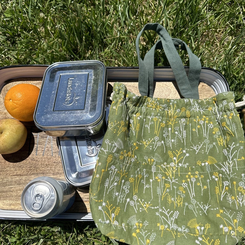 May include: A green reusable tote bag with a white floral pattern. The bag is sitting on a wooden surface with a stainless steel lunchbox, a can of soda, and a piece of fruit.