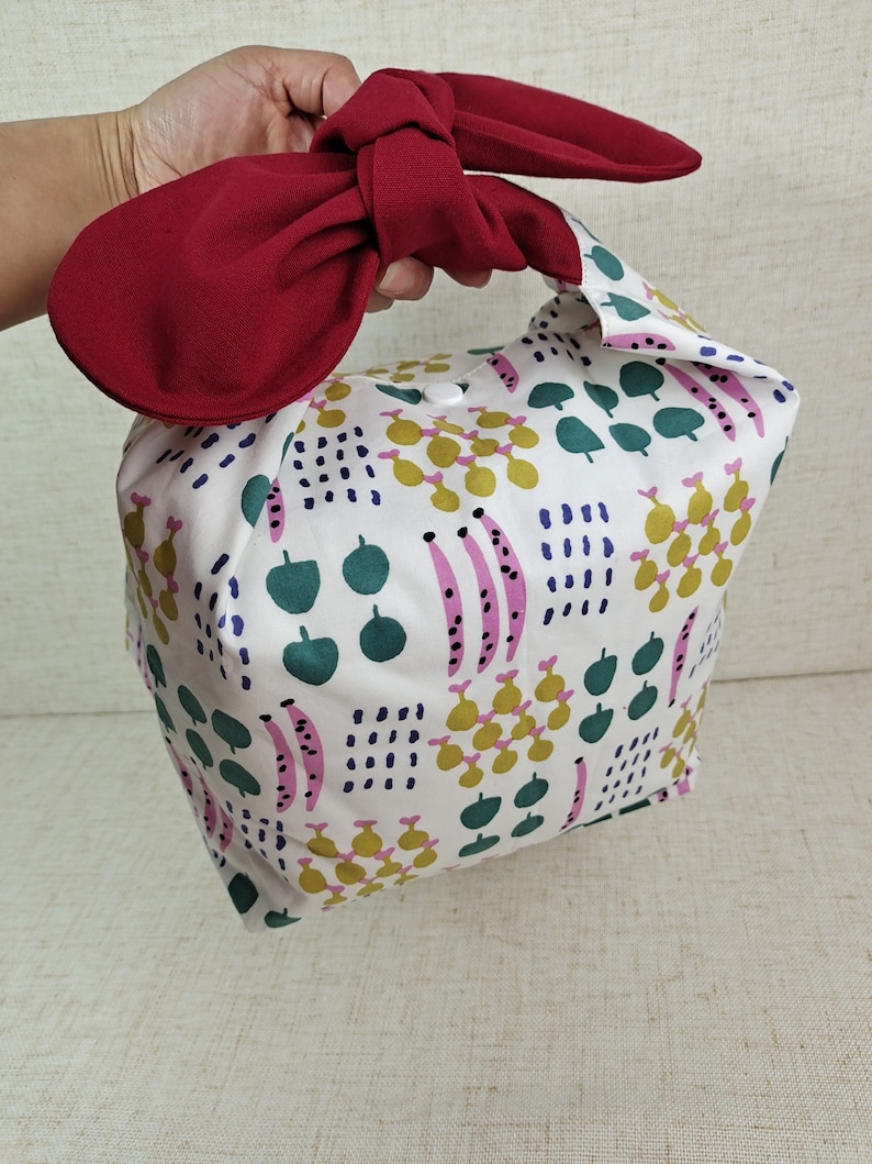 May include: A white fabric tote bag with a red bow on the top. The bag has a colorful geometric pattern with green, pink, yellow, and blue accents.