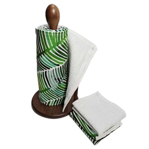 Pu&ograve; includere: Un set di asciugamani da cucina riutilizzabili con un motivo a foglie tropicali verdi e bianche. Gli asciugamani sono arrotolati su un supporto in legno e ci sono tre asciugamani extra impilati su un asciugamano verde e bianco.