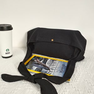 May include: Black canvas tote bag with a knotted strap and a gold button closure. The bag is open and a magazine titled "Geographic Secrets of the Whales" is inside.