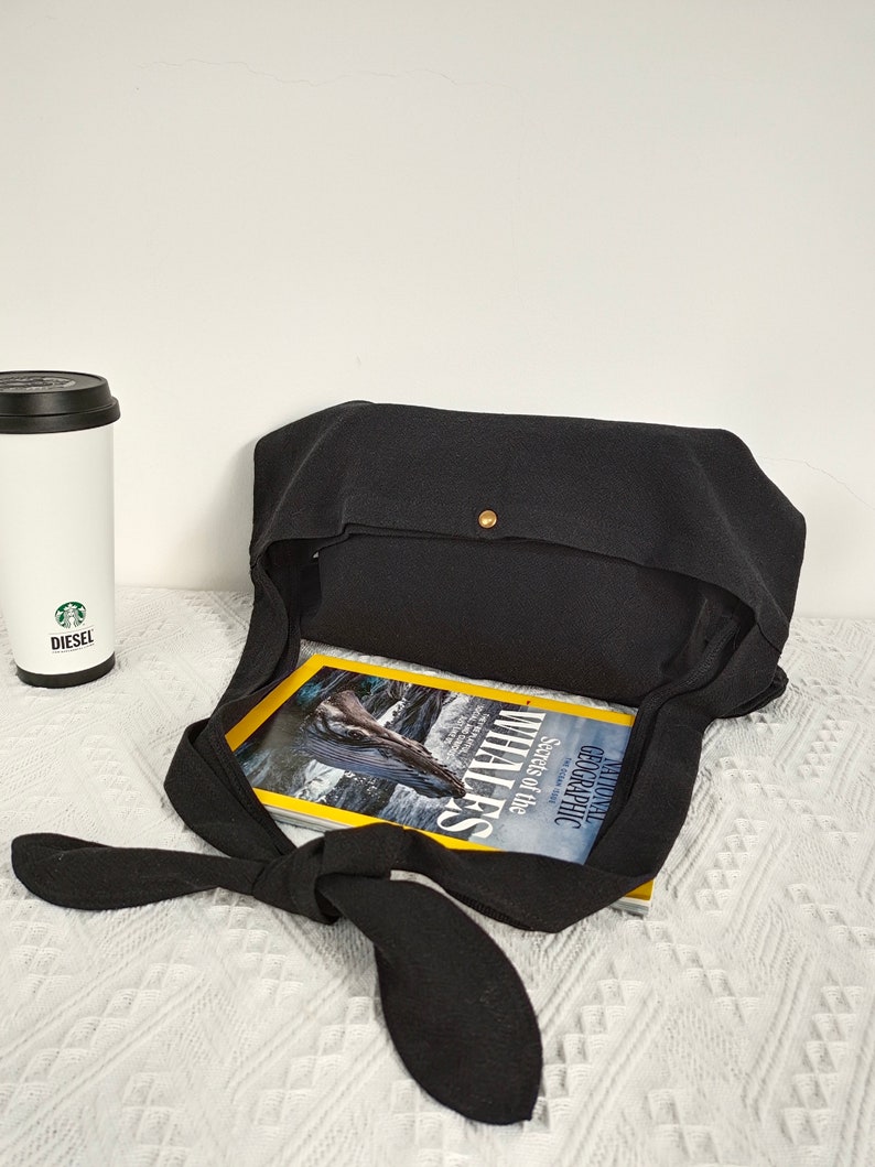 May include: Black canvas tote bag with a knotted strap and a gold button closure. The bag is open and a magazine titled "Geographic Secrets of the Whales" is inside.