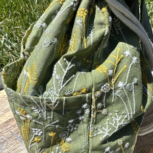 May include: A green linen drawstring bag with white and yellow embroidered floral designs. The bag has a single pocket on the front.