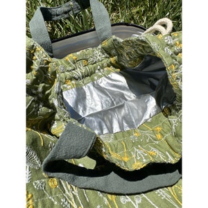May include: Green canvas tote bag with white and yellow floral embroidery. The bag has a silver lining and two green canvas handles.