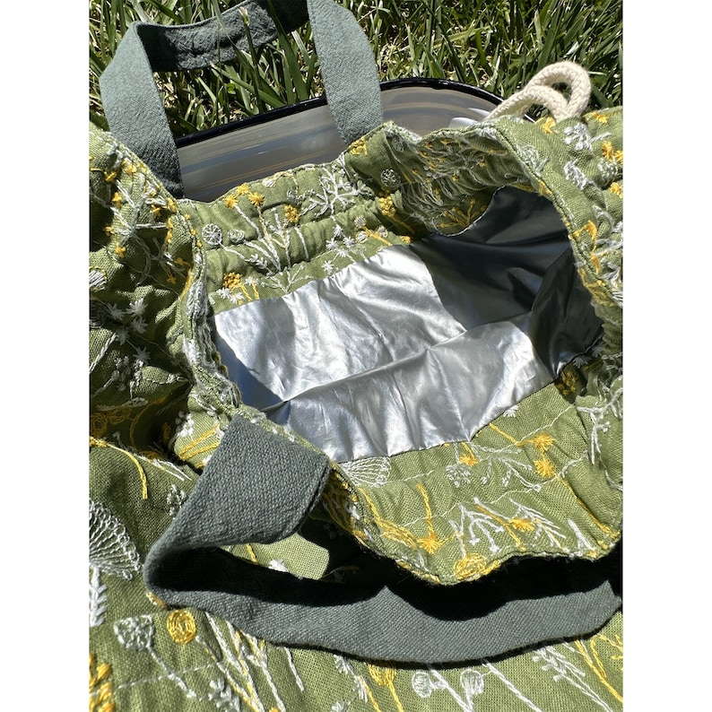 May include: Green canvas tote bag with white and yellow floral embroidery. The bag has a silver lining and two green canvas handles.