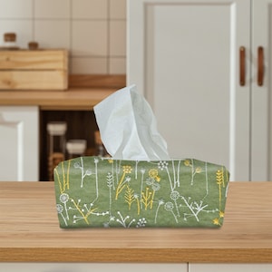 Puede incluir: Una funda para caja de pañuelos de color verde con un patrón floral en blanco y amarillo. La funda tiene una textura de lino y está llena de pañuelos blancos.