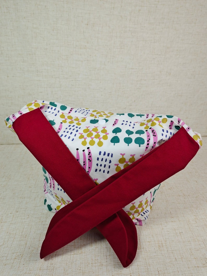May include: A white fabric headband with a colorful abstract pattern and a red bow tie. The pattern includes green, blue, pink, and yellow shapes.