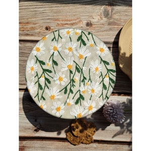 May include: A round, beige fabric coaster with an embroidered design of white daisies with yellow centers and green stems.