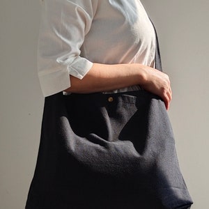 May include: A black canvas tote bag with a single gold button closure. The bag is worn over the shoulder of a person wearing a white shirt.