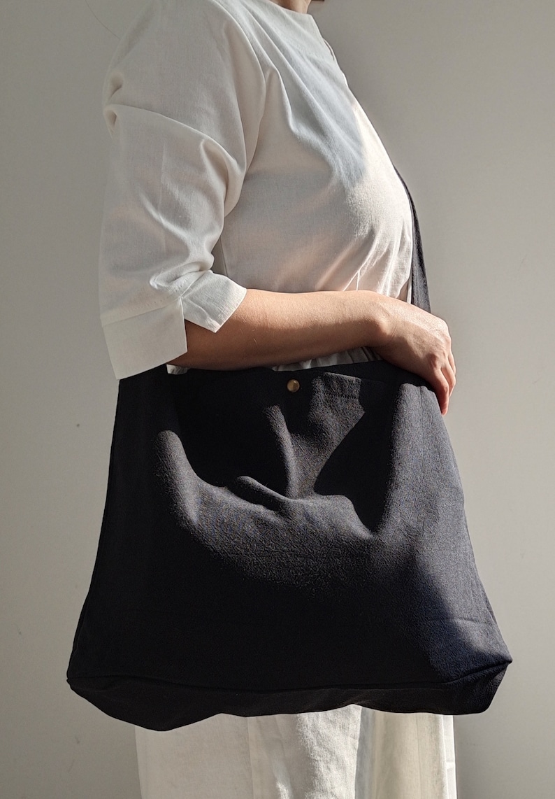 May include: A black canvas tote bag with a single gold button closure. The bag is worn over the shoulder of a person wearing a white shirt.