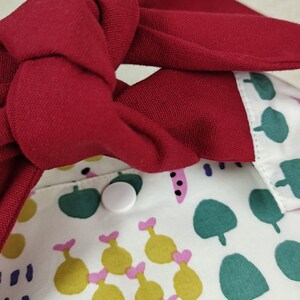 May include: A white fabric bag with a red fabric strap tied in a knot. The bag has a green, yellow, and pink pattern. A white button is visible on the bag.