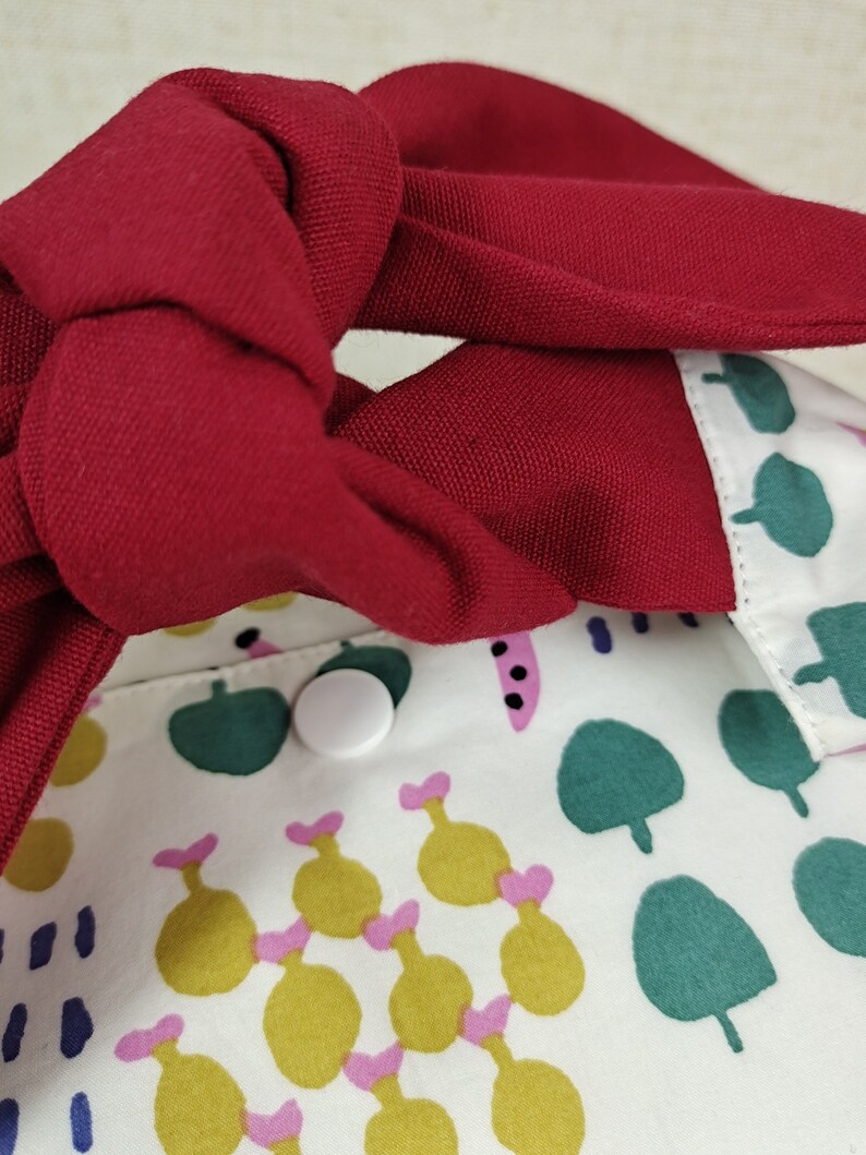 May include: A white fabric bag with a red fabric strap tied in a knot. The bag has a green, yellow, and pink pattern. A white button is visible on the bag.