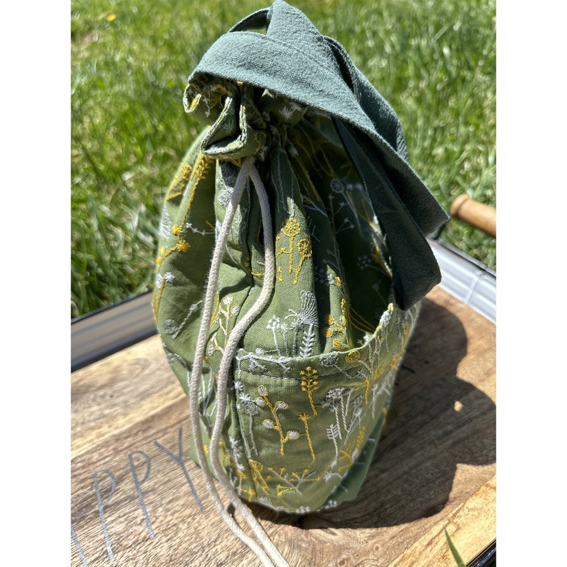 May include: A green drawstring bag with white and yellow floral embroidery. The bag has a long, white drawstring and a green fabric handle.