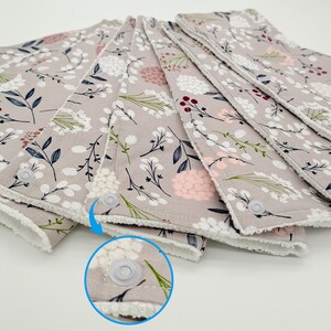 May include: A stack of six gray fabric burp cloths with a white floral pattern and a white snap closure. The cloths have a white fleece backing.