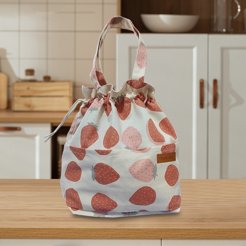 May include: A white canvas drawstring bag with a pink and red strawberry print. The bag has a brown leather tag with the text "Charlie Arts" on it.