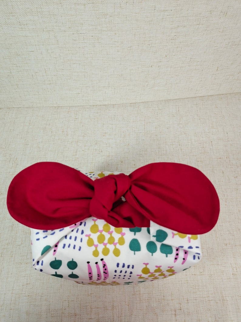 May include: A white fabric gift bag with a colorful geometric pattern, tied with a red ribbon bow.