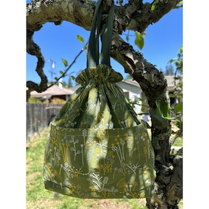 May include: A green drawstring bag with a floral pattern embroidered in white and yellow. The bag is hanging from a tree branch.