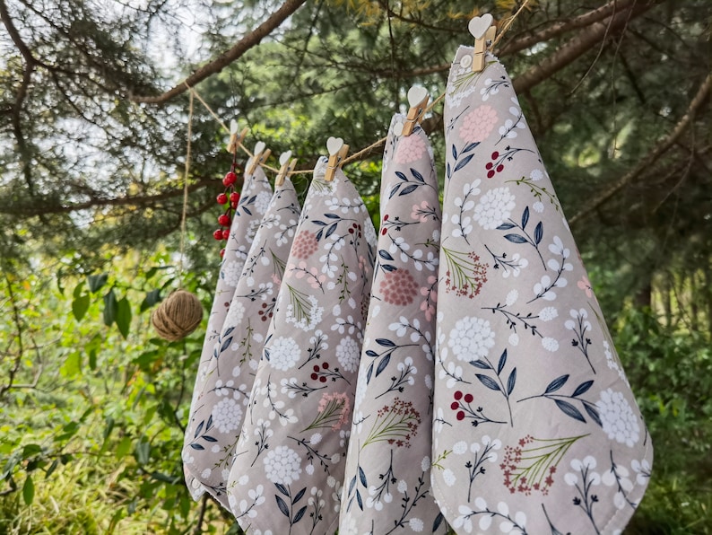 May include: Five tea towels hanging on a clothesline outdoors. The towels are a light gray with a floral pattern in shades of white, pink, and red.