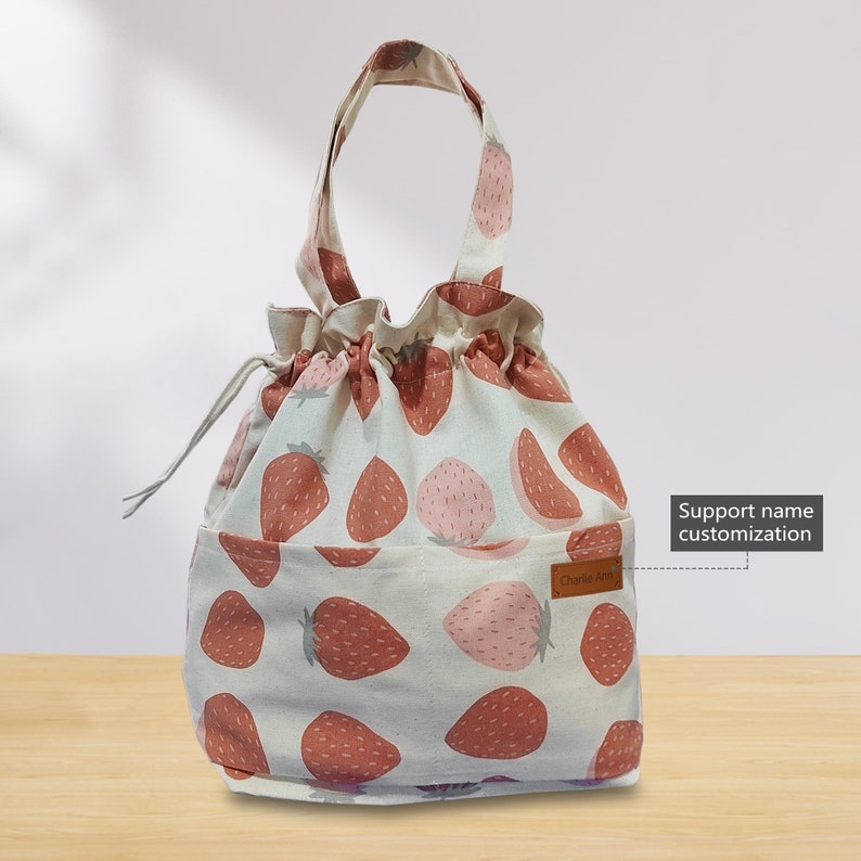 May include: A white drawstring bag with a strawberry print. The bag has a brown leather handle and a brown leather patch with the text "Charlie Art" on it. The bag has a pocket on the front. Support name customization.