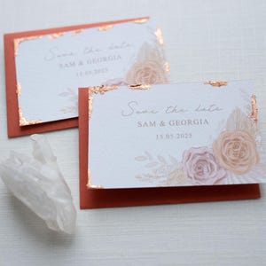 Könnte beinhalten: Save-the-Date-Karte mit einem floralen Design in Rosa- und Pfirsichtönen. Die Karte hat goldene Folienakzente und den Text "Save the date Sam & Georgia 15.05.2025".