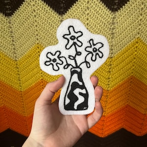 May include: A white felt patch with black embroidery of a vase with three flowers. The vase has a black and white swirl pattern.