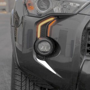 Könnte beinhalten: Nahaufnahme der vorderen Stoßstange eines grauen SUV mit einem schwarzen Kühlergrill, einem klaren Scheinwerfer und einem gelben Blinklicht. Die Stoßstange hat eine weiße Akzentverkleidung.