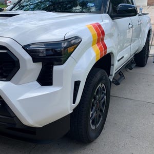 Puede incluir: Camioneta blanca con faros delanteros negros y una calcomanía de rayas rojas, amarillas y blancas en el lateral. La camioneta tiene estribos laterales negros y ruedas negras.