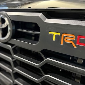 May include: Close-up of a black truck grille with a silver Toyota logo and a red, orange, and yellow "TRD" emblem.