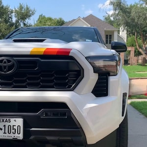 Puede incluir: Camioneta blanca con parrilla negra y faros negros. La camioneta tiene una franja roja, amarilla y naranja en el capó. La camioneta tiene una placa de matrícula de Texas que dice "1050".