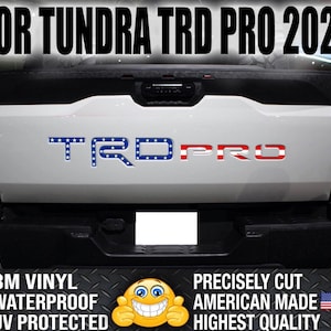 Puede incluir: Un portón trasero de camioneta blanca con un emblema "TRD PRO" rojo, blanco y azul. El emblema tiene estrellas en la parte azul y rayas rojas en la parte roja. El texto "FOR TUNDRA TRD PRO 2022" está en la parte superior de la imagen. El texto "3M VINYL", "WATERPROOF" y "UV PROTECTED" están listados debajo del emblema. El texto "PRECISELY CUT", "AMERICAN MADE" y "HIGHEST QUALITY" están listados debajo del emblema. Un emoji sonriente está a la izquierda del texto. Una bandera estadounidense está a la derecha del texto.