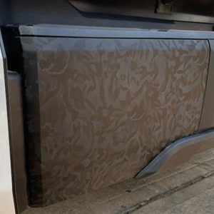 Pode incluir: Close-up do para-choque traseiro de um caminhão com um padrão de camuflagem. O para-choque é cinza escuro com um design texturizado. Uma placa de licença com o número 1239 é visível.