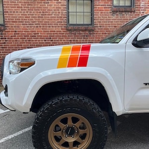 Puede incluir: Camioneta blanca con un conjunto de rayas de carreras naranja, rojo y amarillo en el capó. La camioneta tiene una rueda de color bronce con un neumático negro.