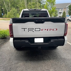 Op de afbeelding: Witte pick-up truck met een zwarte achterklep en een rood "TRD PRO" embleem. De truck heeft een zwarte bumper en een trekhaak.