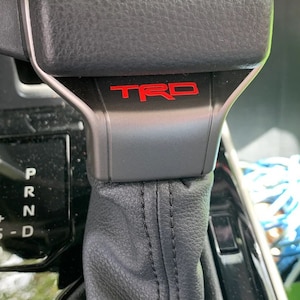 Peut inclure: Gros plan sur un pommeau de levier de vitesses en cuir noir avec une garniture argentée et un logo "TRD" rouge.