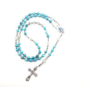 May include: A rosary featuring turquoise-colored beads, clear faceted beads, and silver-toned metal spacers. The beads spell out the name EMMA in silver letter blocks. A silver-toned crucifix hangs at the bottom, and a central oval medal depicts a figure.