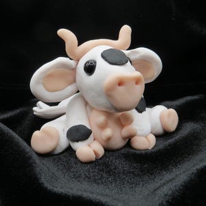 May include: A handmade cow figurine in a sitting position. The cow is primarily white with black spots, pink ears, a pink nose, and small horns. The figurine is set against a black background.