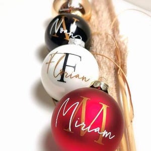 Könnte beinhalten: Personalisierte Weihnachtskugeln in Rot, Weiß, Schwarz und Gold. Jede Kugel trägt einen Namen in einer stilvollen Schriftart. Die Kugeln sind mit dekorativen Trockengräsern angeordnet.