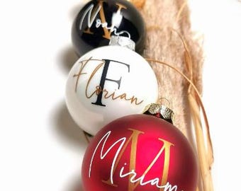 May include: A row of four spherical Christmas ornaments in gold, black, white, and red. Each ornament features a large capital letter and a name printed in a contrasting script font. The names visible are Lisa, Noah, Florian, and Miriam.