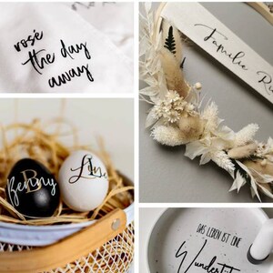 May include: A collection of home decor items with handwritten calligraphy. The items include a white fabric with the text "rose the day away", a white ceramic dish with the text "Das Leben ist eine Wundertite", a white egg with the text "Lisa", a black egg with the text "Benny", a wooden hoop with dried flowers and the text "Familie Ris", and a white wicker basket.