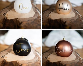 May include: Four Christmas ornaments in white, gold, black, and bronze. Each ornament has a name in a cursive font: Oma, Heidi, Jonas, and Michaela. The ornaments have gold-colored tops and are displayed on a wooden surface.