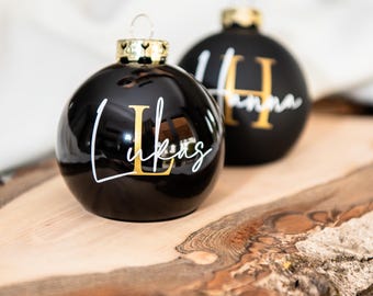 May include: Two round black Christmas ornaments with gold metallic caps. Each ornament features a large gold letter and a white script name printed over it. One ornament displays the name Lukas with the letter L, and the other displays Hanna with the letter H.