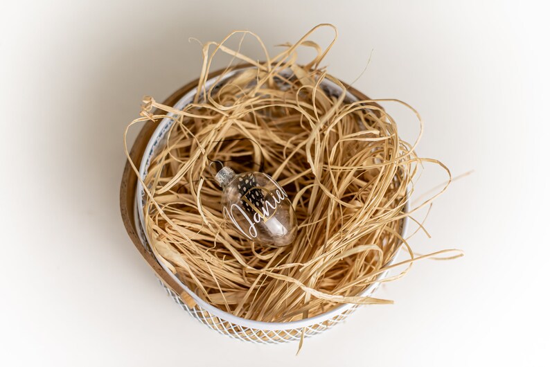 May include: A clear glass ornament with black and white speckled paint and the name "Jamie" written in white calligraphy is nestled in a white wire basket filled with shredded tan paper.