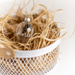 May include: A clear glass Christmas ornament with a gold inscription that reads "Daniel" and a small black and white feather inside. The ornament is sitting in a white wire basket filled with brown shredded paper.