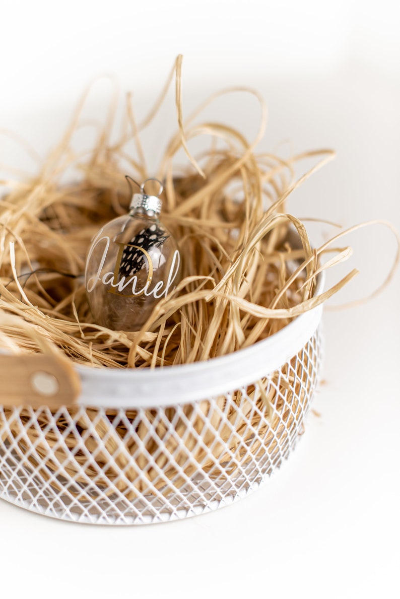 May include: A clear glass Christmas ornament with a gold inscription that reads "Daniel" and a small black and white feather inside. The ornament is sitting in a white wire basket filled with brown shredded paper.