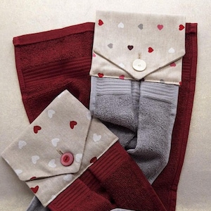 Op de afbeelding: Een set van twee handdoeken, één bordeauxrood en één grijs, met hartvormige stoffen kleppen. De kleppen zijn beige met rode, witte en grijze harten en knoopsluitingen. De handdoeken zijn opgevouwen en gerangschikt.