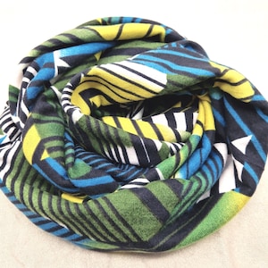 Könnte beinhalten: Gerollter Schal mit einem geometrischen Muster in Grün-, Blau-, Gelb-, Schwarz- und Weißtönen. Der Schal ist aus einem weichen Stoff gefertigt und eng gerollt, wodurch das farbenfrohe Design zur Geltung kommt. Das Muster umfasst Streifen und geometrische Formen.