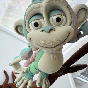 May include: A 3D-printed monkey figurine with a friendly expression, perched on a brown branch. The monkey features a light blue and cream color scheme with accents of green and pink. The figurine has a detailed, layered design.