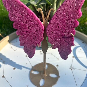 May include: A purple butterfly-shaped decorative object with a textured surface, displayed on a gold metal stand with a round base. The stand is on a white surface with a gold star pattern.
