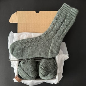 May include: A gray knitted sock with a lacy pattern, displayed in a cardboard box with two balls of gray yarn.
